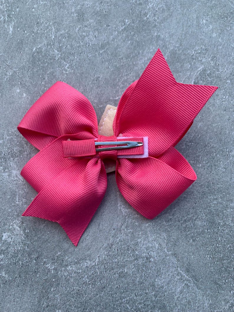 Puede incluir: Un gran lazo rosa con un clip de cocodrilo plateado. El lazo est&aacute; hecho de cinta de grosgrain y est&aacute; atado en un estilo cl&aacute;sico.