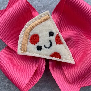 Puede incluir: Un lazo rosa para el pelo con una aplicaci&oacute;n de fieltro en forma de rebanada de pizza. La rebanada de pizza tiene una cara sonriente con pepperoni rojo y una corteza dorada.