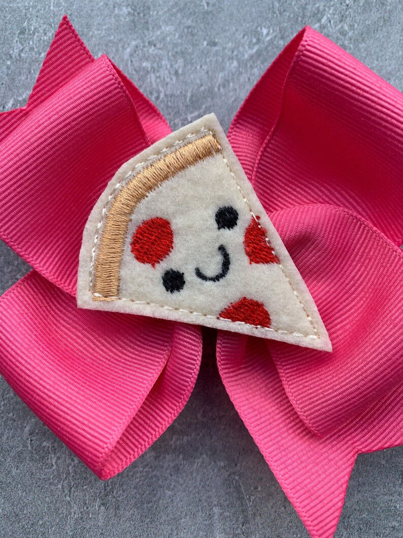 Puede incluir: Un lazo rosa para el pelo con una aplicaci&oacute;n de fieltro en forma de rebanada de pizza. La rebanada de pizza tiene una cara sonriente con pepperoni rojo y una corteza dorada.