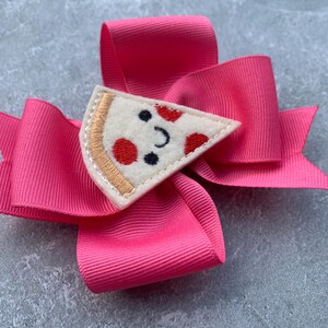 Puede incluir: Un lazo rosa para el pelo con una aplicaci&oacute;n de fieltro en forma de rebanada de pizza. La rebanada de pizza tiene una cara sonriente con pepperoni rojo y una aceituna negra.