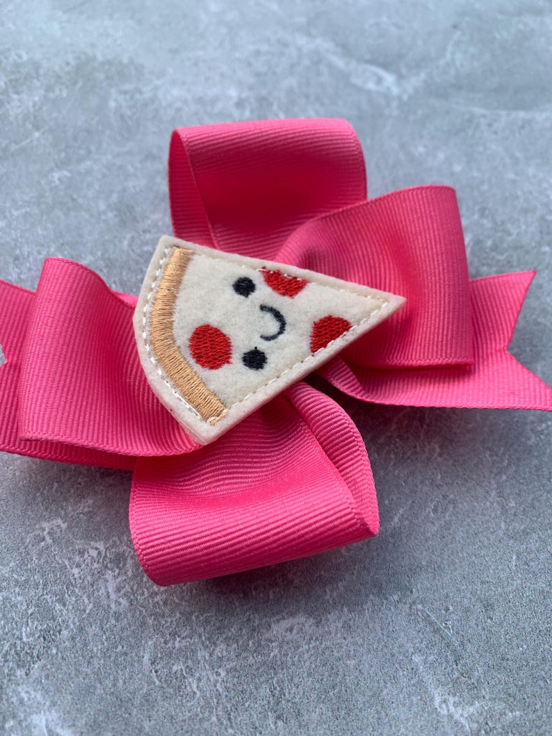 Puede incluir: Un lazo rosa para el pelo con una aplicaci&oacute;n de fieltro en forma de rebanada de pizza. La rebanada de pizza tiene una cara sonriente con pepperoni rojo y una aceituna negra.