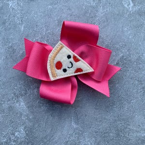 Puede incluir: Un lazo rosa con una aplicaci&oacute;n de fieltro en forma de rebanada de pizza. La rebanada de pizza tiene pepperoni rojo, una corteza blanca y una cara sonriente.