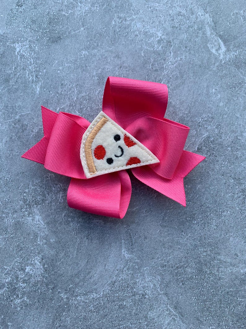 Puede incluir: Un lazo rosa con una aplicaci&oacute;n de fieltro en forma de rebanada de pizza. La rebanada de pizza tiene pepperoni rojo, una corteza blanca y una cara sonriente.