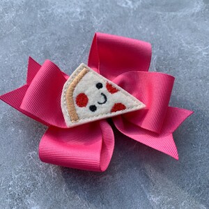 Puede incluir: Un lazo de tela rosa con una aplicaci&oacute;n de fieltro en forma de rebanada de pizza. La rebanada de pizza tiene pepperoni rojo y una cara sonriente.