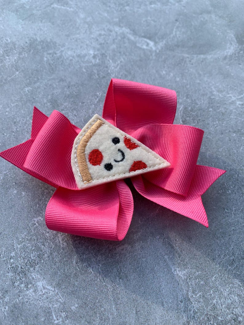 Puede incluir: Un lazo de tela rosa con una aplicaci&oacute;n de fieltro en forma de rebanada de pizza. La rebanada de pizza tiene pepperoni rojo y una cara sonriente.