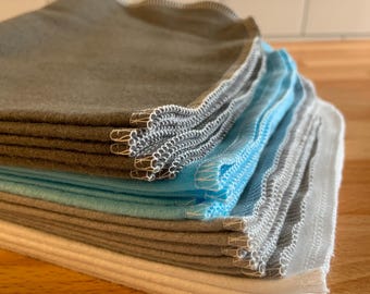 May include: A stack of folded, square cloths in various colors: gray, light blue, and white. Each cloth has a serged edge with white thread. The cloths are stacked on a wooden surface, with a white tile backsplash in the background.