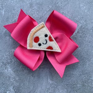 Puede incluir: Un lazo rosa para el pelo con una aplicaci&oacute;n de fieltro en forma de rebanada de pizza. La rebanada de pizza tiene pepperoni rojo, una corteza blanca y una cara sonriente.