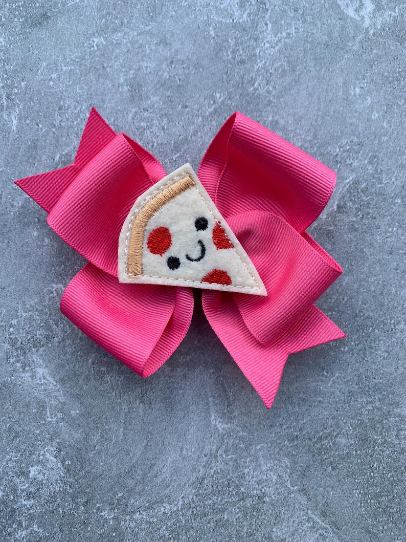 Puede incluir: Un lazo rosa para el pelo con una aplicaci&oacute;n de fieltro en forma de rebanada de pizza. La rebanada de pizza tiene pepperoni rojo, una corteza blanca y una cara sonriente.
