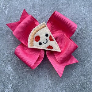 Puede incluir: Un lazo rosa para el pelo con una aplicaci&oacute;n de fieltro en forma de rebanada de pizza. La rebanada de pizza tiene pepperoni rojo, una corteza blanca y una cara sonriente.