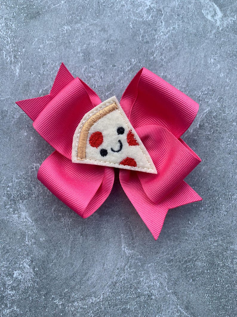 Puede incluir: Un lazo rosa para el pelo con una aplicaci&oacute;n de fieltro en forma de rebanada de pizza. La rebanada de pizza tiene pepperoni rojo, una corteza blanca y una cara sonriente.
