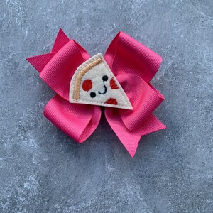 Puede incluir: Un lazo rosa con una aplicaci&oacute;n de fieltro en forma de rebanada de pizza. La rebanada de pizza tiene una cara sonriente y pepperoni rojo.
