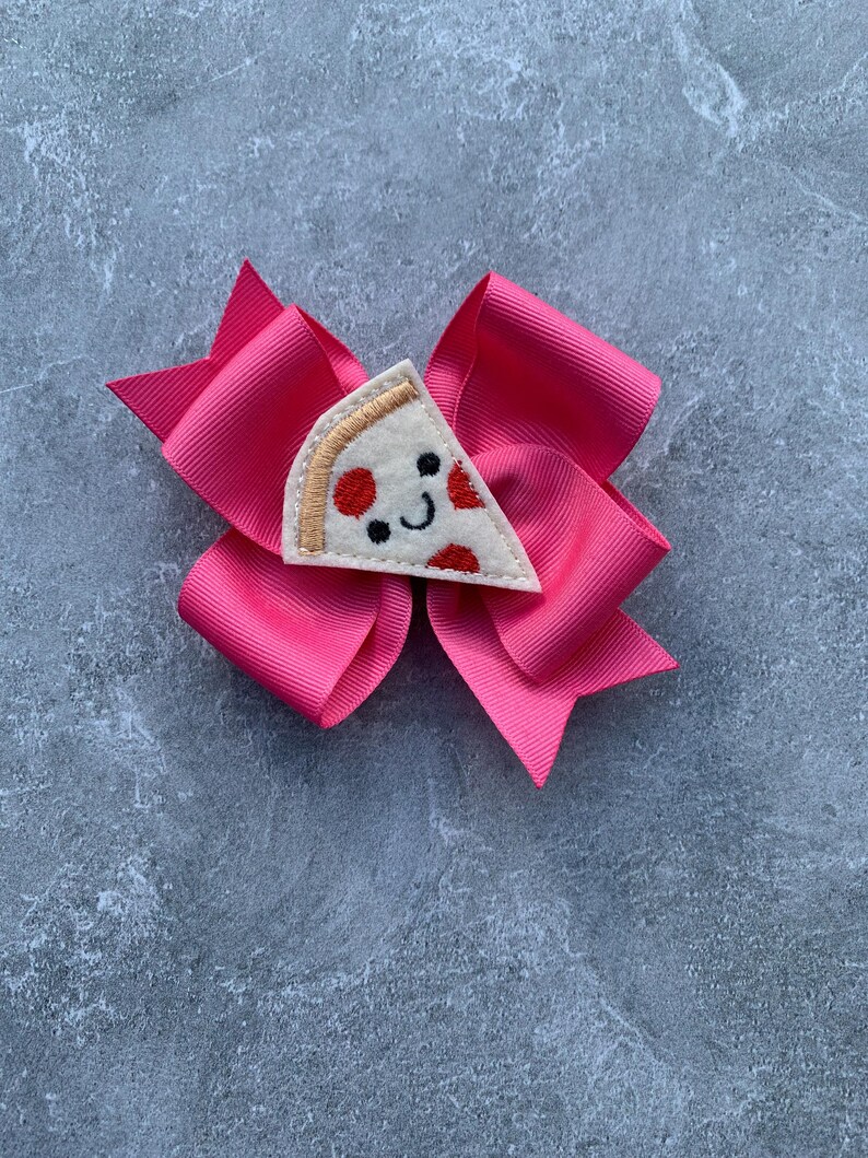 Puede incluir: Un lazo rosa con una aplicaci&oacute;n de fieltro en forma de rebanada de pizza. La rebanada de pizza tiene una cara sonriente y pepperoni rojo.