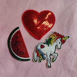 Puede incluir: Tres parches para planchar: un corazón rojo, una rodaja de sandía y un unicornio arcoíris. Los parches están hechos de lentejuelas y bordados.