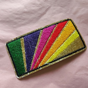 May include: A colorful embroidered patch with a rainbow design. The patch has a gold glitter background and is outlined in black.