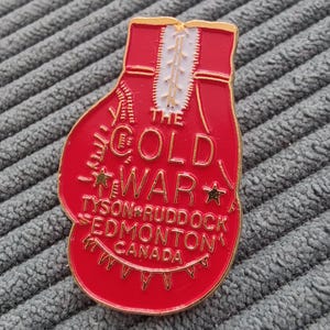 May include: A red and gold enamel pin in the shape of a boxing glove. The pin features the text "THE COLD WAR" and "TYSON RUDDOCK EDMONTON CANADA". The glove has a white zipper detail.