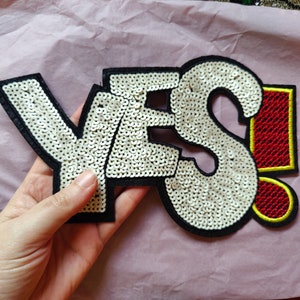 Può includere: Una toppa bianca con paillettes con la parola "YES!" in lettere nere bordate. Il punto esclamativo è rosso con un contorno giallo.