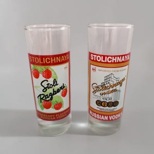 以下が含まれることがあります： 赤と白の「STOLICHNAYA」の文字が入った透明なショットグラスが2つ。1つのグラスにはラズベリーのイラストと「Stoli Razberi」が、もう1つには「IMPORTED FROM RUSSIA」と「RUSSIAN VODKA」の文字が入っています。
