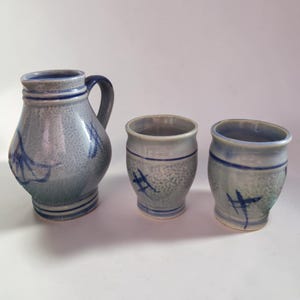 May include: A ceramic pitcher and two matching cups. The set is a muted blue-gray with darker blue accents and abstract painted designs. The pitcher has a handle and a rounded body. The cups are cylindrical.