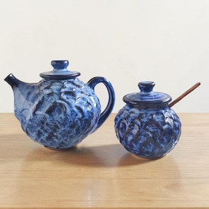 May include: A blue ceramic teapot and a small sugar bowl with a wooden spoon. Both items have a textured surface and a dark blue glaze. The teapot has a handle and a spout. The sugar bowl has a lid.