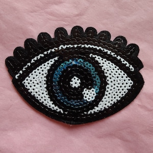 May include: A black and white eye patch with a blue sequin eye. The eye patch is made of fabric and has a scalloped edge.