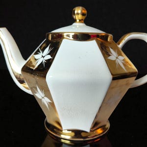 Könnte beinhalten: Weiß-goldene Teekanne mit geometrischem Design. Die Teekanne hat einen weißen Körper mit goldenen Akzenten und einen goldenen Deckel. Weiße florale Designs befinden sich auf den goldenen Abschnitten. Griff und Ausguss sind weiß mit goldenen Verzierungen.