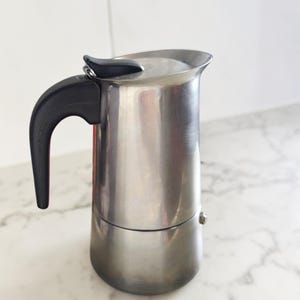 Può includere: Una caffettiera in acciaio inossidabile con manico e coperchio neri. La caffettiera è cilindrica con la parte superiore arrotondata e un manico nero. La caffettiera è appoggiata su un piano di lavoro in marmo bianco.