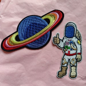 May include: Two embroidered iron-on patches. One is a colorful planet with rings. The other is a cartoon astronaut giving a thumbs up.