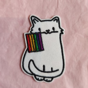 Könnte beinhalten: Weißer gestickter Bügelaufkleber mit einer Katze, die eine Regenbogenflagge hält. Die Katze ist schwarz umrandet und hat ein einfaches Gesicht.