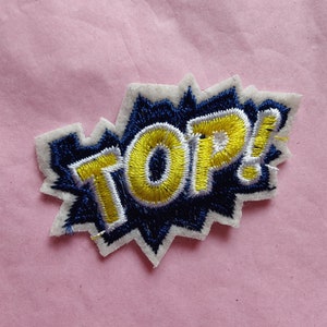 May include: A white, blue, and yellow embroidered patch with the word "TOP!" in a comic book style font.