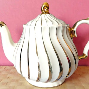 Może przedstawiać: Biały ceramiczny imbryk ze złotymi akcentami. Imbryk posiada pionowe złote paski oraz złoty uchwyt, dzióbek i gałkę pokrywki. Design jest klasyczny, elegancki i nadaje się do serwowania herbaty.