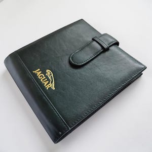 May include: Dark green leather notebook with a gold Jaguar logo and text. The notebook has a strap closure and a stitched border. The cover is smooth and the overall design is classic and elegant.
