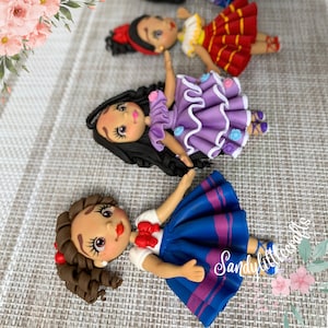 May include: Handmade doll figurines with detailed features. The dolls have dark hair, painted faces, and are dressed in colourful outfits, including a blue skirt, purple dress, and red dress. The dolls are lying on a textured surface.