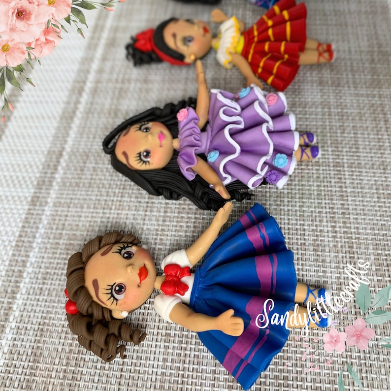 May include: Handmade doll figurines with detailed features. The dolls have dark hair, painted faces, and are dressed in colourful outfits, including a blue skirt, purple dress, and red dress. The dolls are lying on a textured surface.