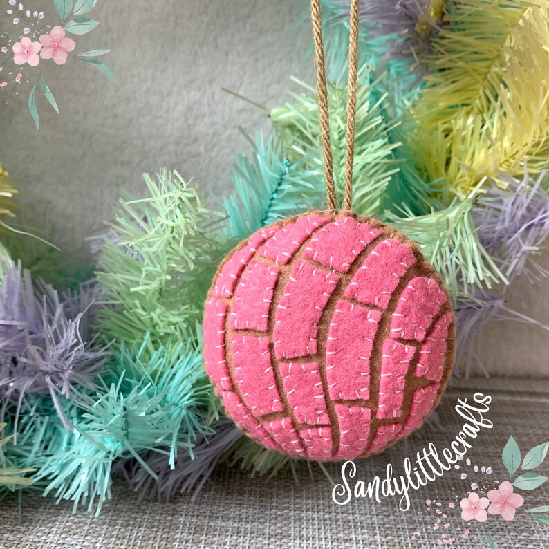 May include: A pink felt ornament shaped like a concha, a traditional Mexican sweet bread. The ornament has a brown base with pink top and white stitched details. It hangs from a natural twine loop, set against a pastel-colored wreath.