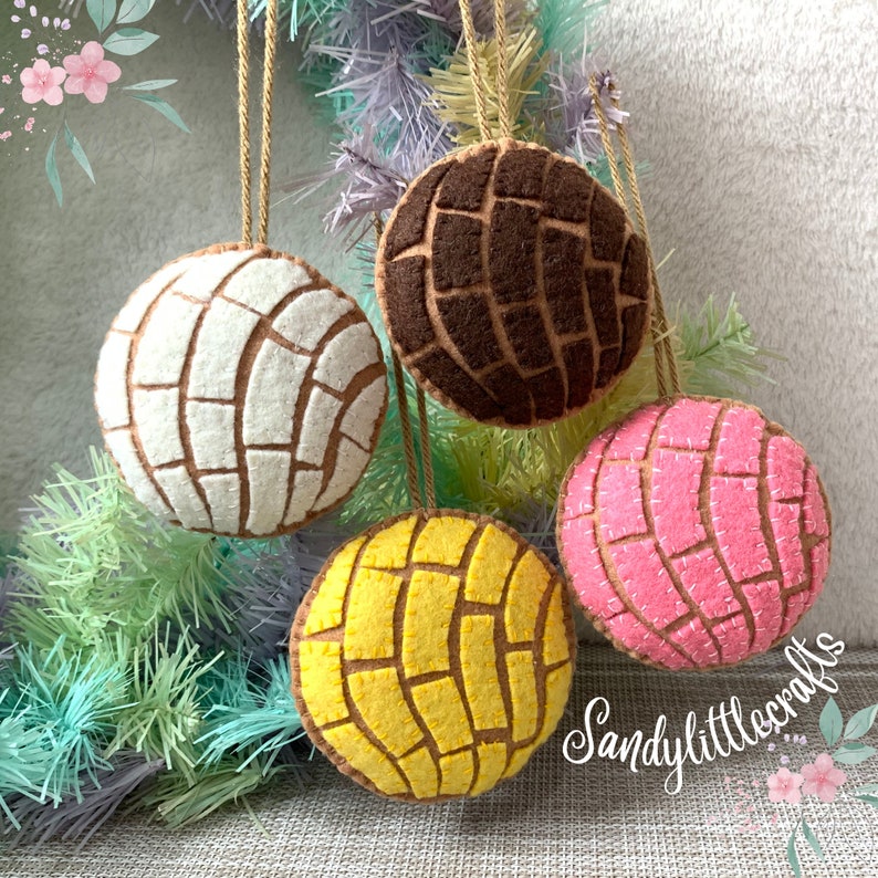 May include: Four felt ornaments shaped like conchas, a traditional Mexican sweet bread. The ornaments are in white, brown, yellow, and pink, each with a brown grid pattern. They hang from brown twine.