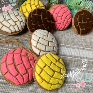May include: A collection of colorful felt ornaments shaped like conchas, a traditional Mexican sweet bread. The ornaments feature various colors, including white, yellow, pink, and brown, with intricate patterns. Each ornament has a brown border and a string for hanging.