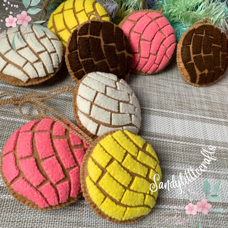 May include: A collection of colorful felt ornaments shaped like conchas, a traditional Mexican sweet bread. The ornaments feature various colors, including white, yellow, pink, and brown, with intricate patterns. Each ornament has a brown border and a string for hanging.