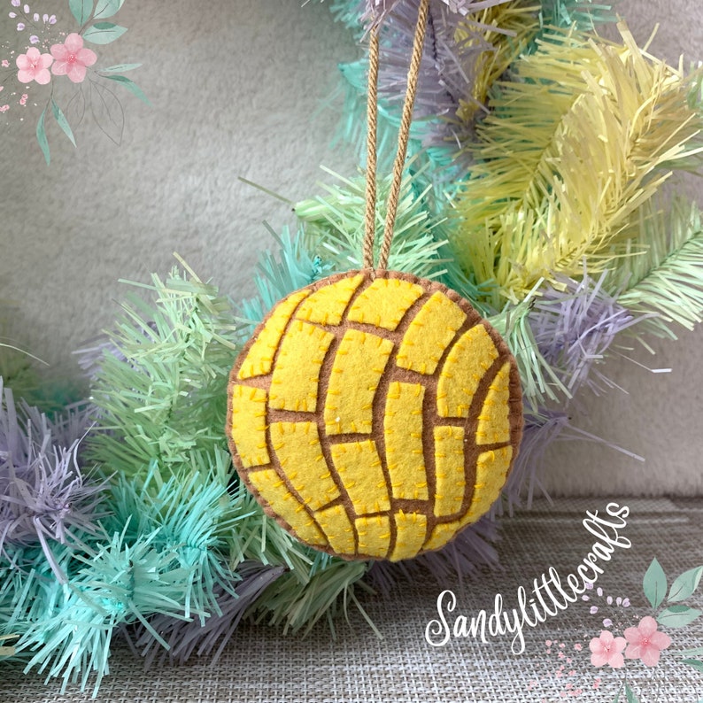 May include: A felt ornament shaped like a concha, a traditional Mexican sweet bread. The ornament is yellow with a brown pattern, and has a gold string for hanging. The ornament is displayed on a pastel-colored wreath.