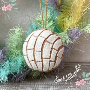 May include: A round, felt ornament resembling a concha pastry, featuring a white and brown pattern. It hangs from a tan string. The background includes pastel-colored decorative branches and the text "Sandylittlecrafts".
