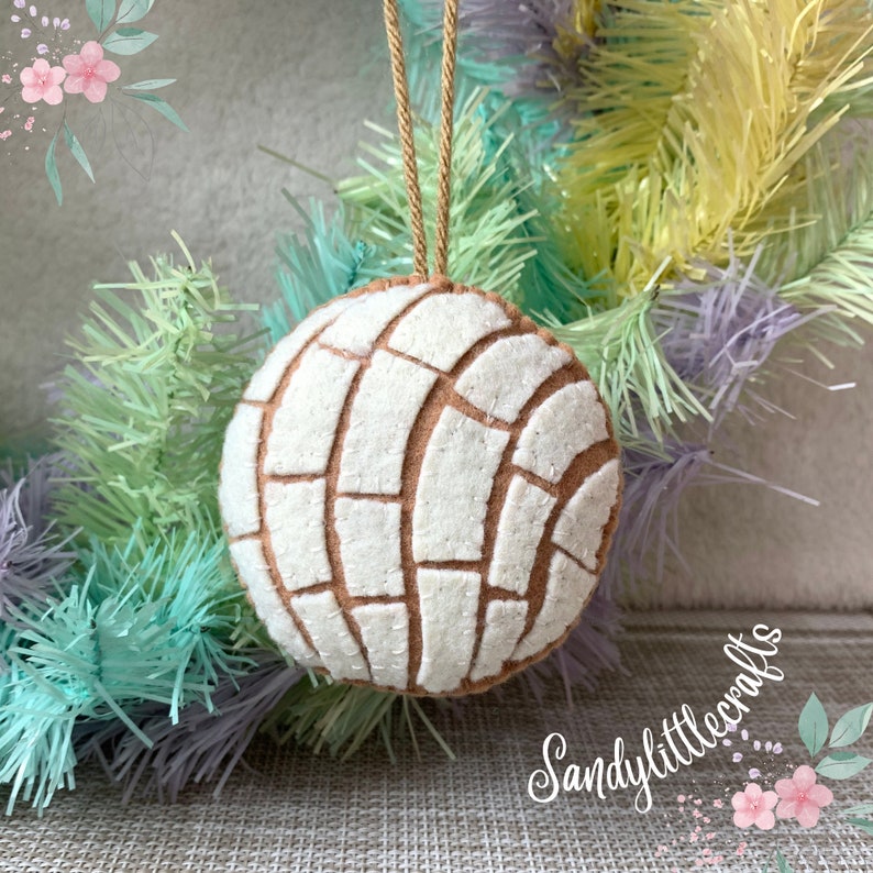 May include: A round, felt ornament resembling a concha pastry, featuring a white and brown pattern. It hangs from a tan string. The background includes pastel-colored decorative branches and the text "Sandylittlecrafts".