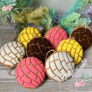 May include: A collection of felt ornaments shaped like conchas, a traditional Mexican sweet bread. The ornaments come in various colors, including yellow, pink, brown, and white, with a textured pattern on top. Each ornament has a loop for hanging.