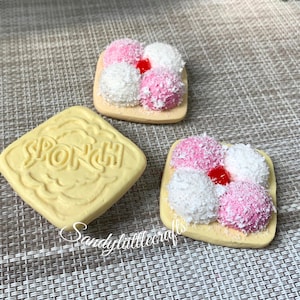 May include: Three miniature square biscuit replicas. One plain light yellow biscuit features the embossed text SPONCH. Two other biscuits are topped with alternating pink and white textured mounds, finished with a small red dot in the centre.