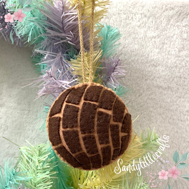 May include: A felt ornament shaped like a concha, a traditional Mexican sweet bread. The ornament is brown with a darker brown grid pattern on top, suspended by a gold string. The background includes a pastel-colored wreath.