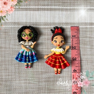 May include: Two handmade polymer clay figurines of girls, one with curly hair and glasses, the other with a red bow. The first wears a blue dress, the second a red dress. A measuring tape is next to the figurines.