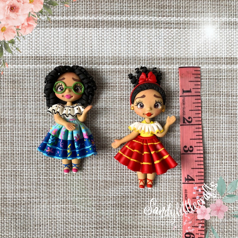 May include: Two handmade polymer clay figurines of girls, one with curly hair and glasses, the other with a red bow. The first wears a blue dress, the second a red dress. A measuring tape is next to the figurines.