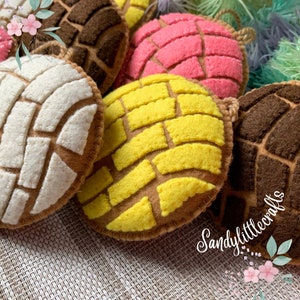 May include: Close-up of several colorful felt concha bread ornaments. The ornaments feature a brown base with white, yellow, pink, and brown patterned tops. Each ornament has a loop for hanging. The image is well-lit and shows the details of the handmade crafts.
