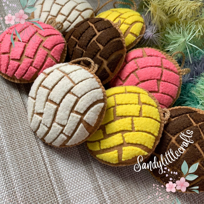 May include: A collection of colorful felt concha ornaments in various colors, including pink, white, yellow, and brown. Each ornament features a detailed, textured design and a loop for hanging. The Sandy Little Crafts logo is visible.