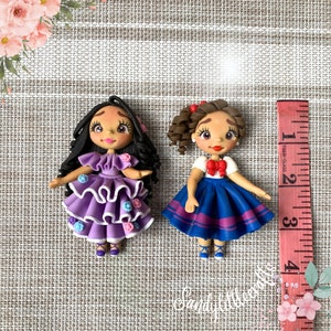 May include: Two handmade polymer clay dolls. One doll wears a purple ruffled dress with floral accents. The other doll is in a white top, blue skirt, and red bow. A ruler shows the dolls are approximately 7.6 cm tall.