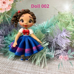 May include: A handmade doll with brown curly hair, red details, and a blue skirt with pink stripes. The doll wears a white top with a red bow. The text "Doll 002" is visible. The doll is a Sandy Little Crafts creation.