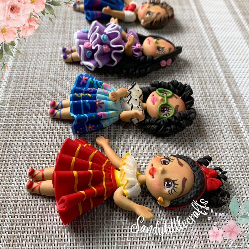May include: Handmade polymer clay figurines of women in colourful dresses. The dolls have detailed facial features, including painted eyes and lips. The dresses are in various colours, including red, blue, and purple, with decorative accents. The dolls are lying down.